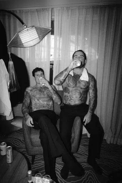 Groom getting ready in a hotel room, shirtless with a towel over his shoulder, beer cans nearby and suit jacket hanging by the window