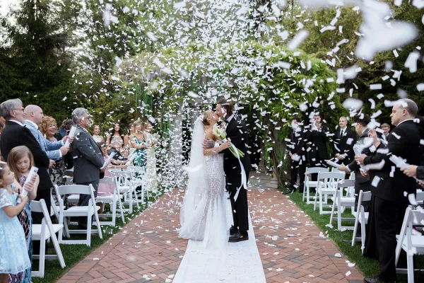 Ceremony kiss moment as bride and groom kiss on a white aisle runner while guests toss confetti in a garden setting