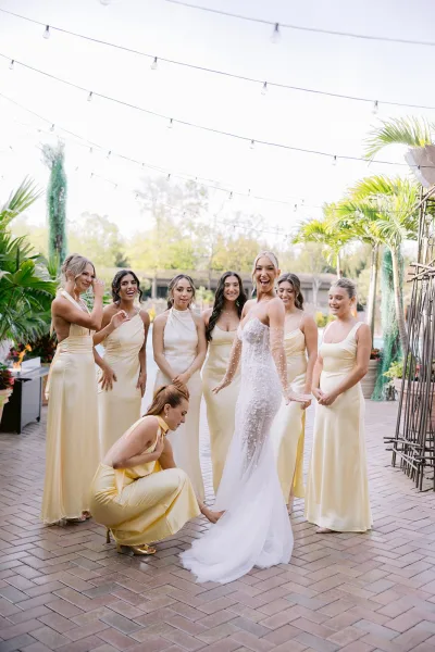 Bride and bridesmaids in champagne bridesmaid dresses help adjust her long veil on a brick patio under string lights with palm trees behind