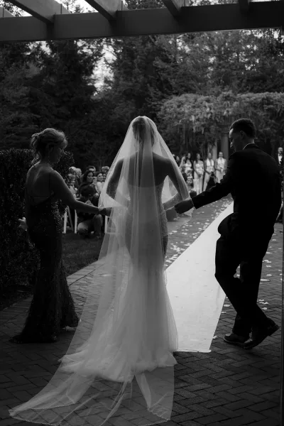 Wedding processional as the bride walks down aisle from behind, long veil trailing over rose petals on a brick garden walkway with guests seated