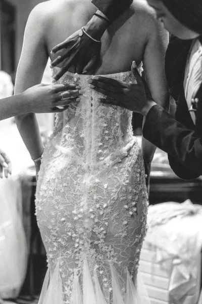 Wedding dress back with button back wedding dress details as hands fasten a beaded corset and bracelets gleam in a softly lit getting-ready room