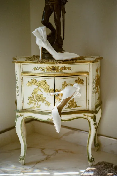 Wedding shoes and bridal heels with ankle-strap bows on a painted side table beside a jewelry chain, lit by sun shadows on marble floor