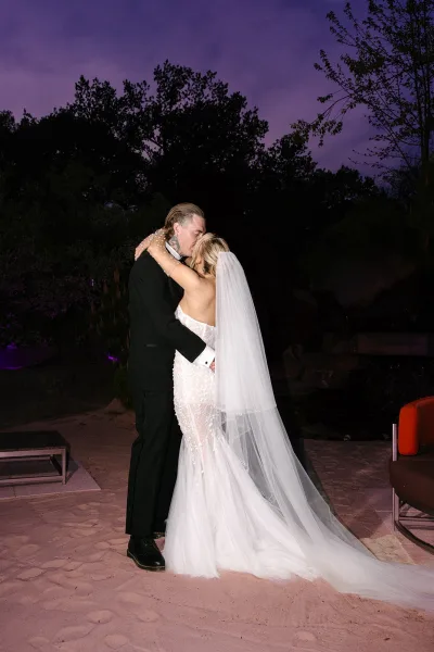 Wedding couple portrait sharing a kiss, bride in a long veil and lace dress embracing groom in tuxedo on an outdoor patio at twilight