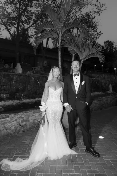 Couple portrait in a black and white wedding portrait style, bride in strapless lace gown with veil holding bouquet beside groom in tux by a stone waterfall at night