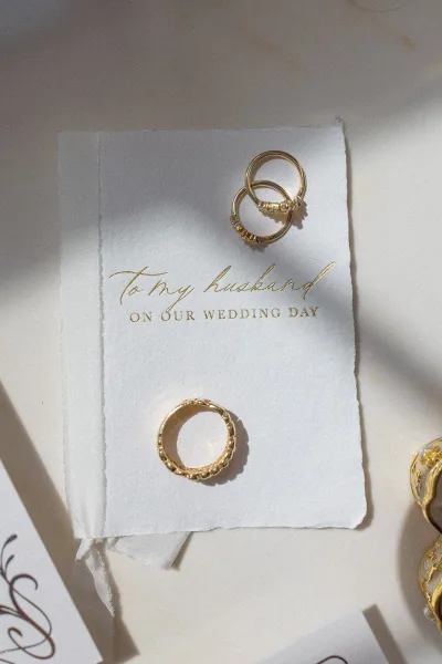 Wedding rings and gold wedding bands stacked on deckled-edge stationery with gold foil calligraphy and a wax seal on a white surface