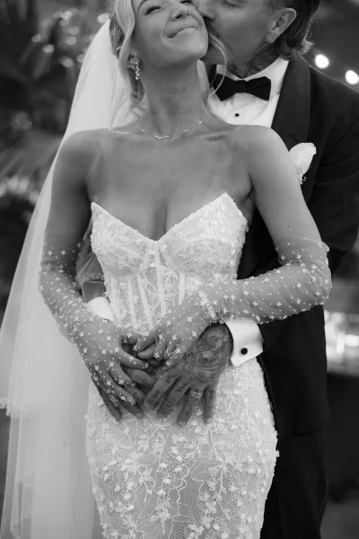 Wedding couple portrait with groom kissing bride’s cheek as they embrace under outdoor greenery and string lights, her veil and lace dress visible