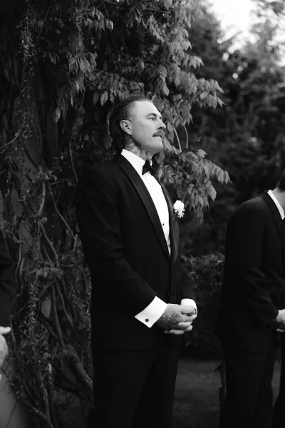 Groom portrait in a black and white wedding portrait style, wearing a black tuxedo with bow tie and boutonniere amid garden foliage