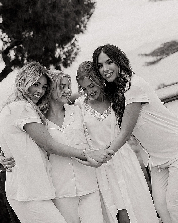 Bridesmaids getting ready in matching bridal party pajamas with lace trim chemise, holding hands on an outdoor terrace by trees and sky