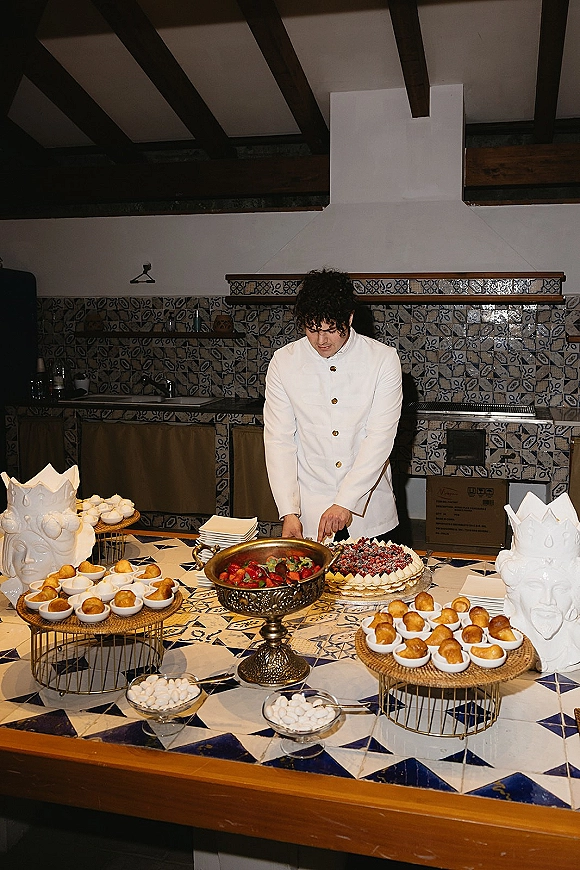 Dessert table with tiered dessert stands, berry-topped cake, meringue cookies and fruit bowl on a blue-and-white tiled counter kitchen backdrop
