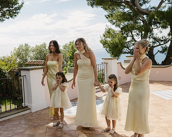 Bridesmaid reaction during bridesmaids first look, laughing on a seaside terrace with champagne dresses, flower girls, and ocean view behind