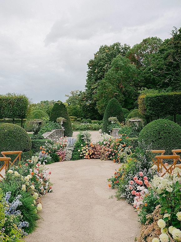 Ceremony aisle decor with outdoor ceremony aisle flowers lining a gravel path, colorful ground arrangements beside wooden chairs in a formal garden
