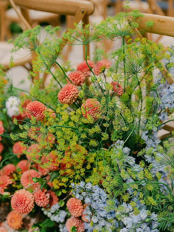 Wedding florals on a ceremony chair florals arrangement with orange blooms, yellow dill flowers, blue delphinium, and greenery outdoors