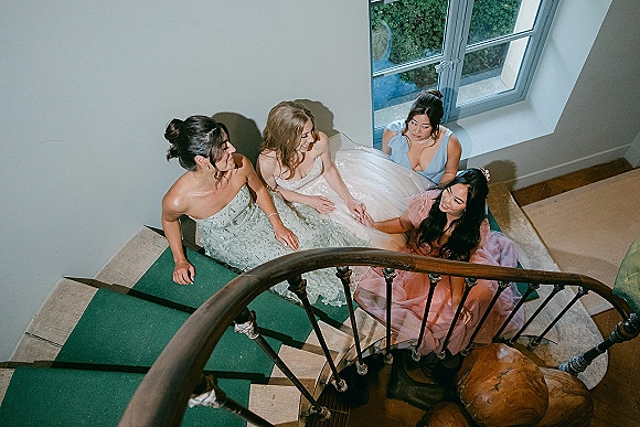 Bride with bridesmaids in a bridal party portrait on an indoor staircase, showing her strapless gown and tiara against white walls and window light