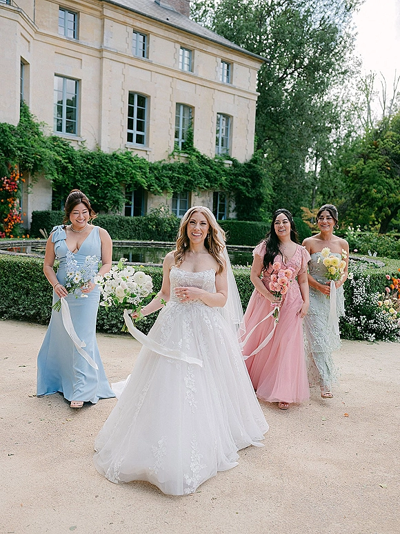 Bride with bridesmaids in pastel dresses walking through an estate garden, holding bouquets with ribbon streamers beside a reflecting pool