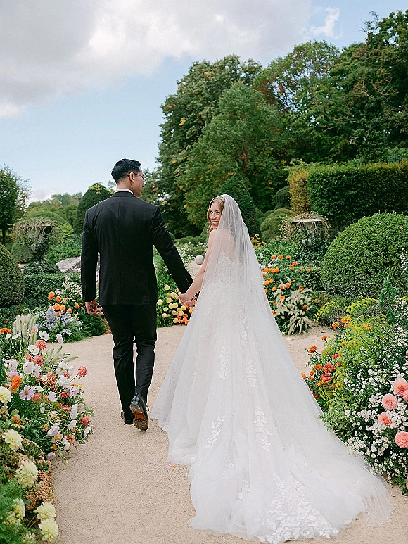 Couple portrait of bride and groom walking away holding hands, bride looking back as cathedral veil trails on a garden path