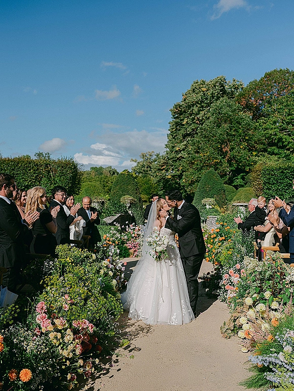 Ceremony kiss as bride and groom embrace on a flower-lined outdoor garden aisle, veil and bouquet visible, guests clapping under blue sky