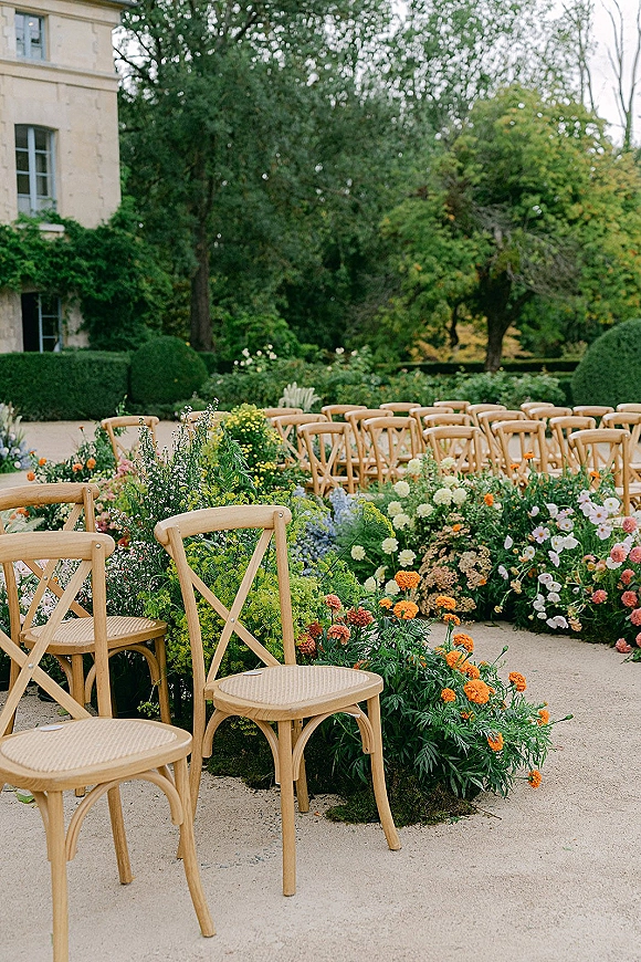 Ceremony aisle decor with wildflower-lined borders beside wood cross-back chairs in a garden courtyard with gravel path and stone exterior