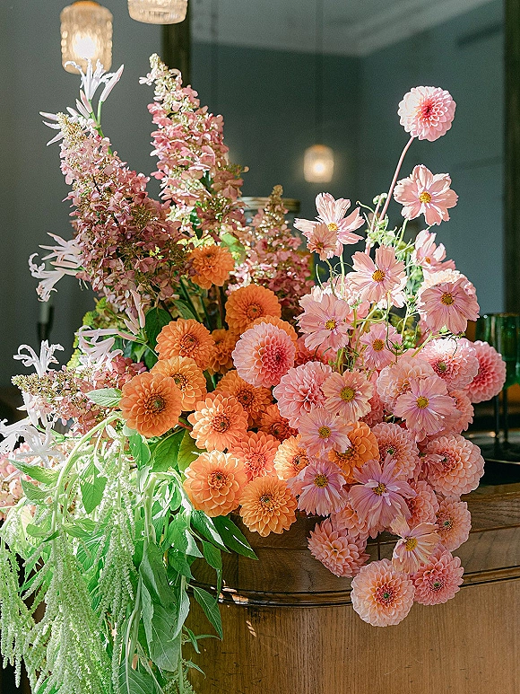 Wedding floral arrangement with dahlias and cosmos in pink and orange atop a wooden bar, pendant lights glowing against a dark wall