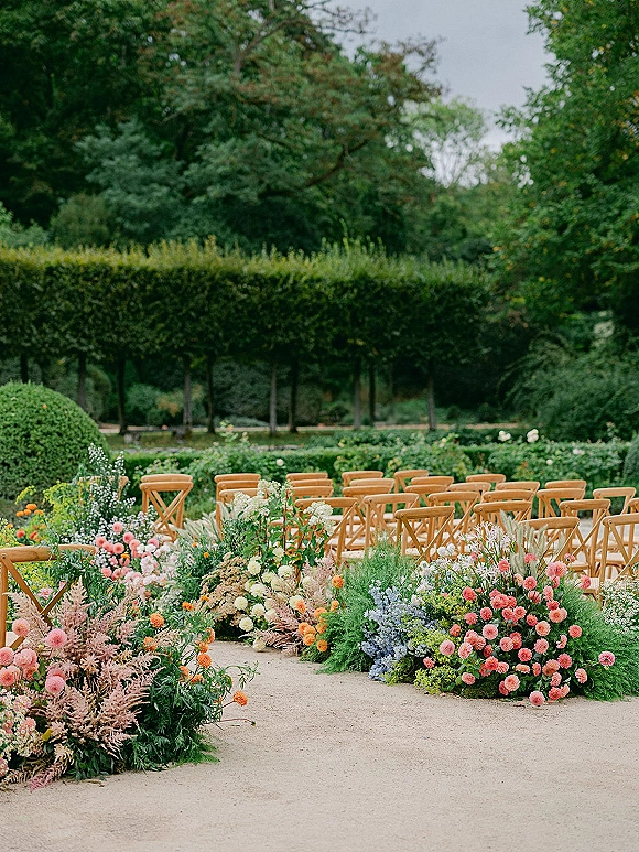 Ceremony aisle decor with outdoor ceremony aisle flowers and greenery beside wood cross back chairs on a gravel garden path under trees