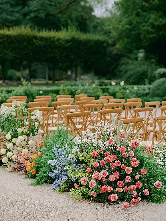 Ceremony aisle decor with outdoor ceremony seating, wooden cross back chairs, and low floral ground arrangements along a gravel path in a garden setting