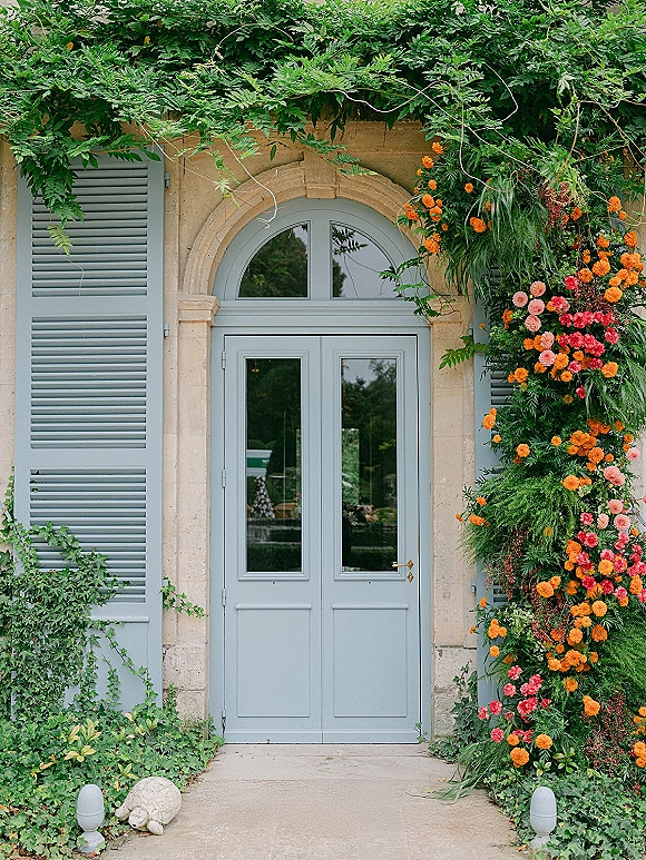 Wedding entrance decor with a floral doorway arch of orange and pink blooms and greenery garland framing shuttered double doors on stone facade