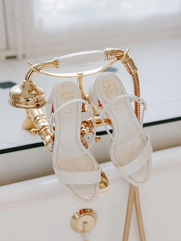 Bridal shoes, white ankle strap heels on a white bathtub beside a gold faucet and handheld shower hose in a tiled bathroom