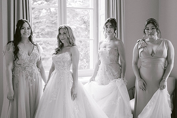 Bridal party portrait of bride with bridesmaids in lace and tulle gowns by a large window, natural light highlighting their jewelry