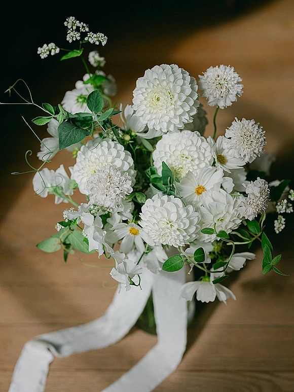 Bridal bouquet of white flowers and greenery in a glass vase, finished with a white ribbon, resting on a wooden table