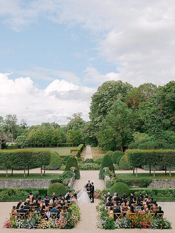 Outdoor wedding ceremony with garden wedding ceremony florals lining a symmetrical aisle, guests seated on crossback chairs in a manicured garden.