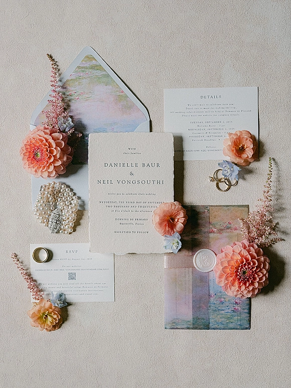 Invitation suite flat lay with deckled edge wedding invitation, RSVP card, wax seal, rings and peach dahlias on neutral textured surface