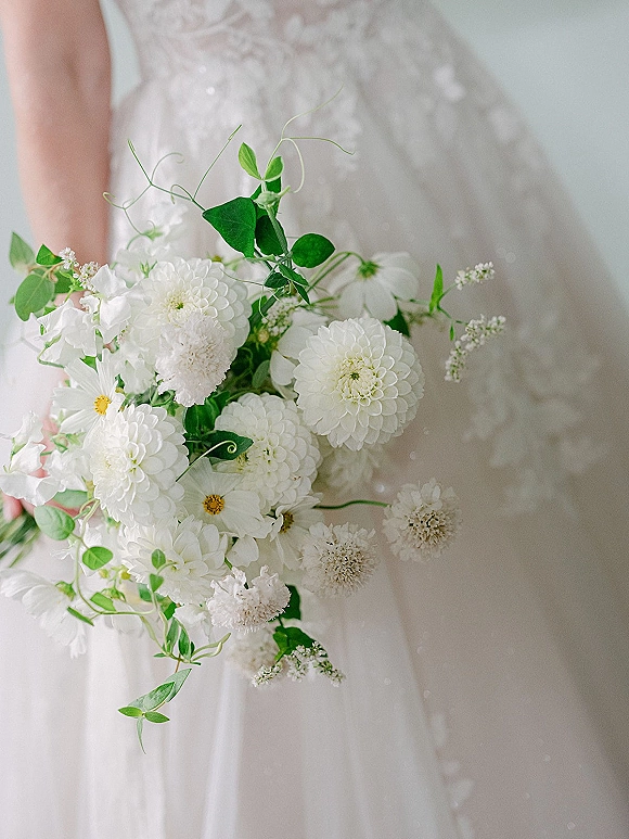 Bridal bouquet of white dahlias and cosmos with greenery and vine tendrils, held against a lace appliqué bridal gown on a soft neutral backdrop