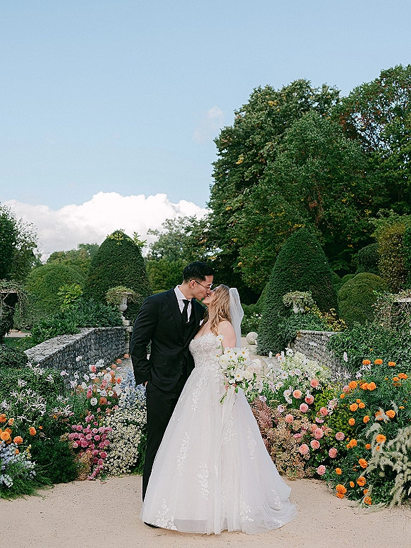 Wedding kiss portrait of bride and groom kiss beside a stone garden bridge, bride in veil holding a daisy bouquet among blooms