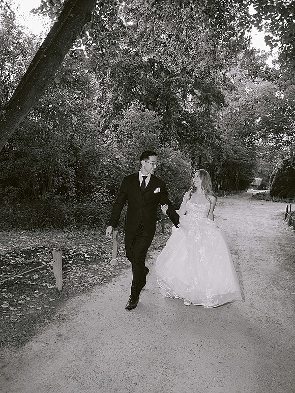 Couple portrait of bride and groom walking arm in arm, bride lifting her gown beside a rope-fenced, tree-lined forest path