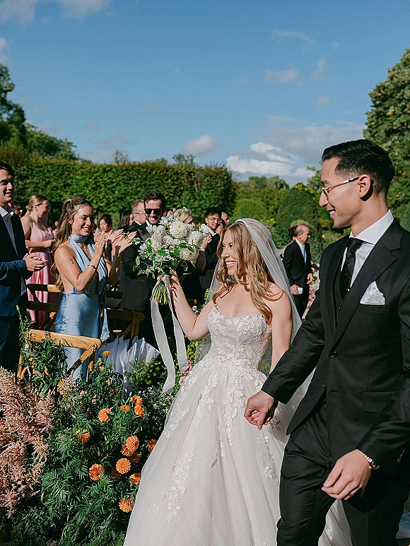 Recessional moment as bride and groom walk the aisle, bride raising a white bouquet with ribbon streamers, guests cheering in a garden setting