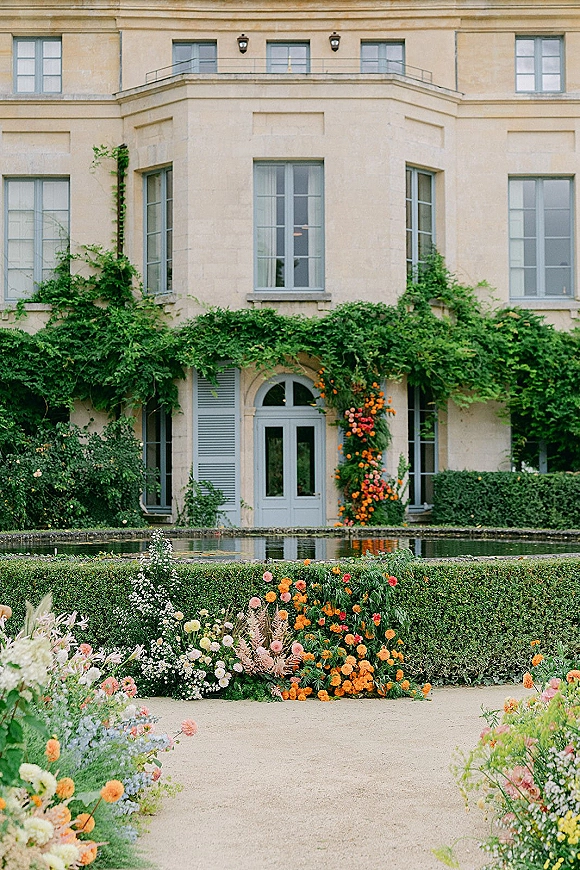 Wedding floral installation with garden flowers and climbing vines framing French doors by a reflecting pool at a stone estate courtyard