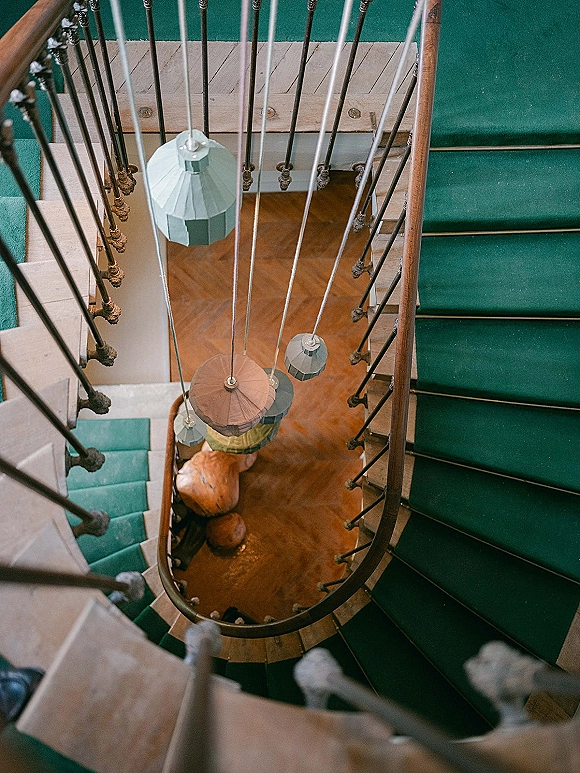 Spiral staircase at a wedding venue with teal stair runner, wrought iron railing, wooden handrail, and hanging pendant lights overhead