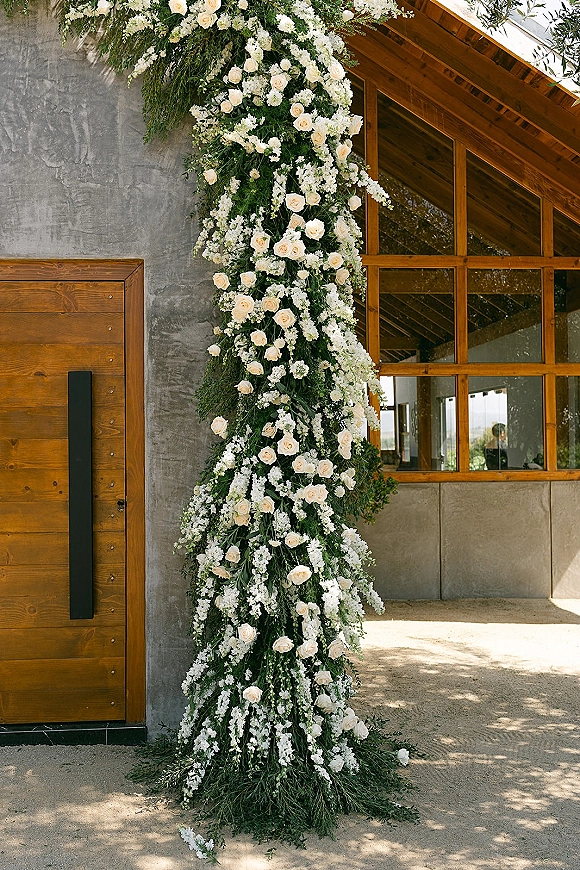 Floral installation of wedding ceremony flowers with cascading white roses and greenery on a pillar outside a modern building entrance walkway