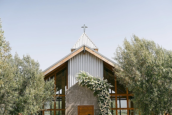 Wedding ceremony backdrop at a chapel wedding ceremony with an asymmetrical rose and greenery garland framing the cross and glass windows under blue sky