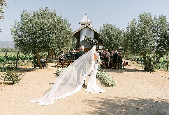 Wedding processional with bride walking down aisle in a lace dress and long veil, escorted past floral arrangements at a vineyard chapel