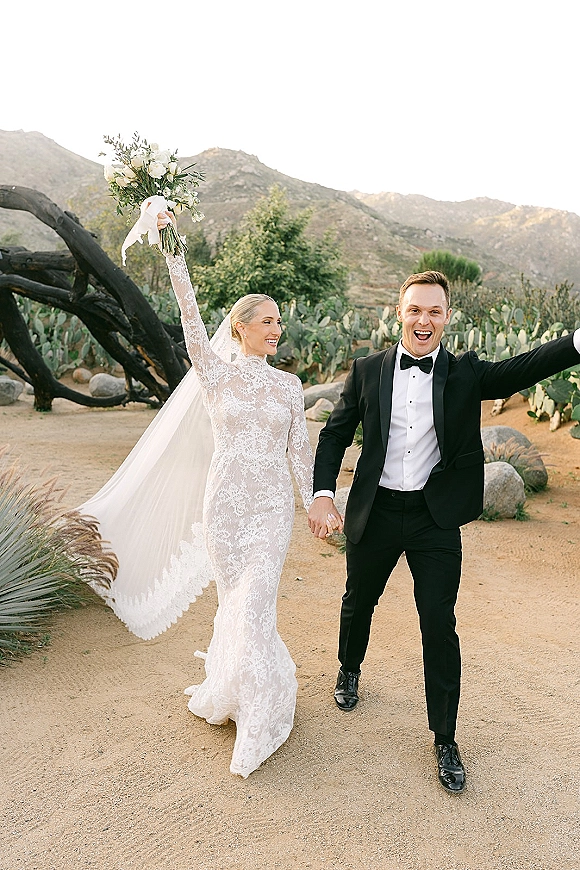 Newlywed couple portrait of bride and groom holding hands, bride in lace long sleeve dress with bouquet, walking through desert cacti and mountains