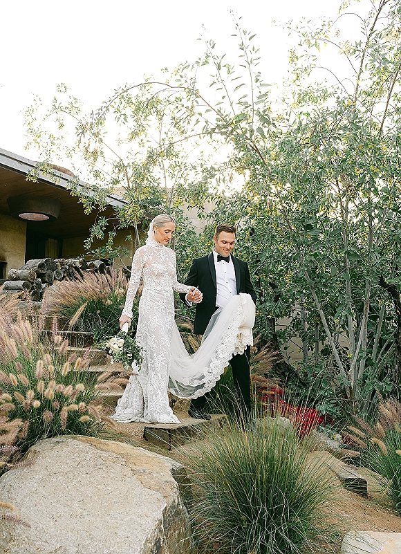Couple portrait of bride and groom holding hands on stone steps, her cathedral veil and lace train flowing in a garden by a modern venue