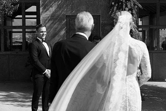 Wedding first look as the groom first look reaction shows him in a black tuxedo while the bride in a lace dress and veil approaches in a sunlit courtyard