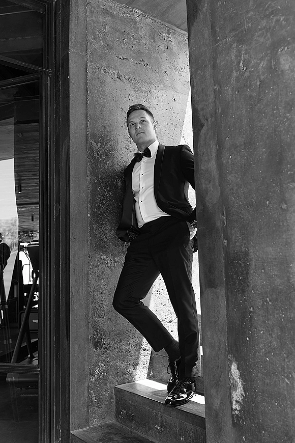 Groom portrait in a black tuxedo with bow tie and wristwatch, leaning by a doorway on steps against a concrete wall backdrop