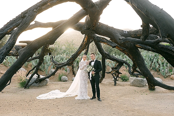 Couple portrait of bride in lace dress and veil with long train holding bouquet beside groom in tuxedo, backlit in desert cactus garden