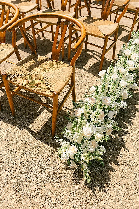 Ceremony aisle decor with ground floral aisle of white roses and greenery between wood chairs, sunlit outdoor setup with soft shadows