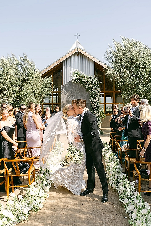 Wedding kiss as bride in lace dress and veil kisses groom in tuxedo, holding bouquet beneath a floral arch outside a chapel with cross