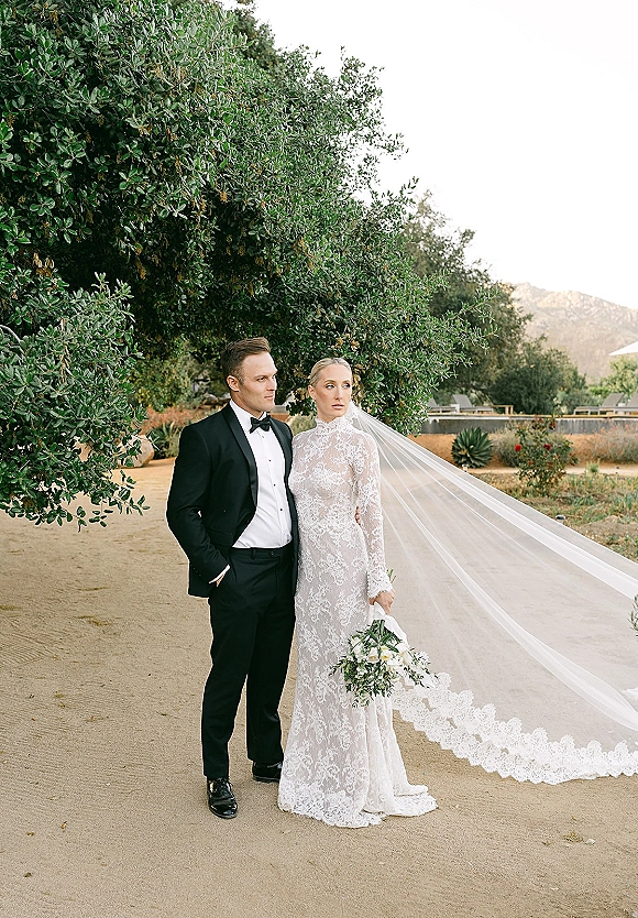 Couple portrait of bride in long sleeve lace dress holding bouquet beside groom in black tuxedo, veil trailing on a garden path with mountains behind