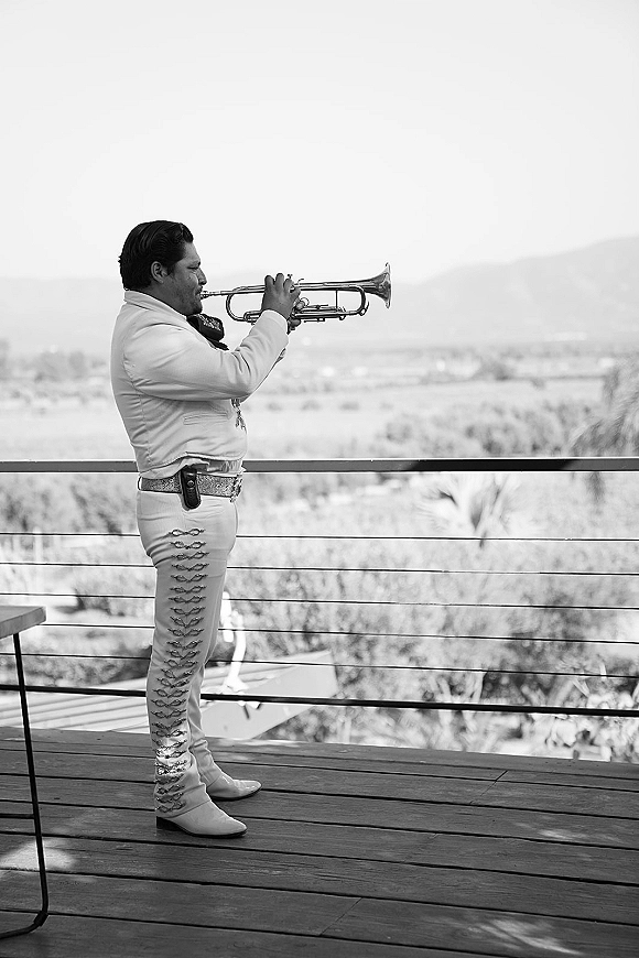 Wedding musician in a mariachi suit playing trumpet on an outdoor terrace, with railing and distant hills and trees behind him