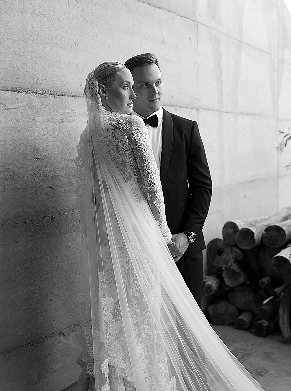 Couple portrait in a black and white wedding portrait, bride looking over shoulder in lace dress and long veil beside groom in tux by concrete wall