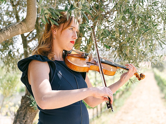 Wedding violinist in a navy dress playing with bow and violin, sunlight filtering through leafy branches along an outdoor path